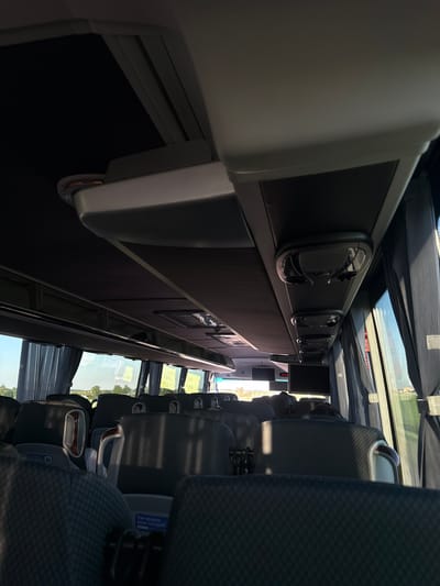 The image shows the interior of a bus, looking forward from the back. The seats are upholstered in a grey patterned fabric, and the overhead compartments are visible, as are the windows and curtains. Outside the windows, a field and trees can be seen under a clear sky. There is a screen mounted on the ceiling towards the front of the bus. There is a blue sticker on one of the seats that reads "Как прошла ваша поездка?" which translates to "How was your trip?". The bus appears to be parked in a rural area, possibly on a road in the vicinity of Абухаўскі сельскі Савет, Belarus, given the landscape.