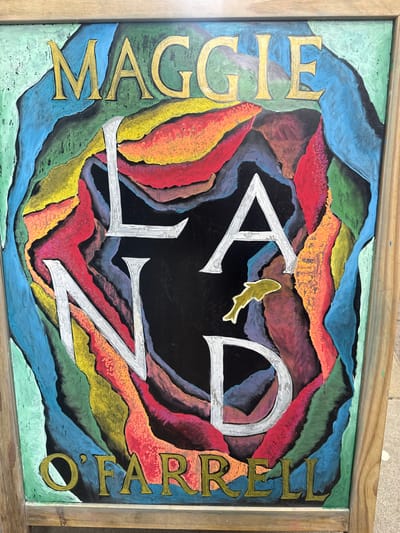 The image shows a colorful chalk drawing on a chalkboard, framed by a light-colored wooden border. The drawing depicts the name "MAGGIE LAND" in large, stylized letters. The letters "MAGGIE" are rendered in gold, while "LAND" is in white with silver outlining. The letters are set against a dark background that is surrounded by swirling, abstract shapes in a variety of vibrant colors including green, blue, red, orange, yellow, and purple. In the center of the "LAND" portion of the drawing, a small, golden fish is depicted swimming. Below the "LAND" text, the name "O'FARRELL" is written in gold letters. The chalkboard is propped up, suggesting it's an outdoor display. The context provided indicates this chalkboard is located in the city of Leominster, United Kingdom.
