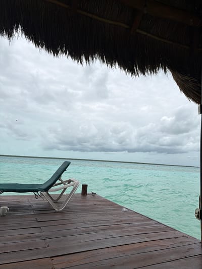 The image depicts a tranquil scene on a wooden dock extending over a body of turquoise water. A green lounge chair is positioned on the dock, facing the water. A white mug sits on the left side of the dock near the chair. The water is calm with slight ripples, and the sky is overcast with dense clouds. In the distance, a thin strip of green vegetation marks the horizon. The foreground features the weathered texture of the wooden planks. The scene is framed by the thatched roof of a structure overhead, casting a shadow over the top portion of the image. The location is identified as Colonia Aarón Merino, Mexico. There are no people visible in the frame.