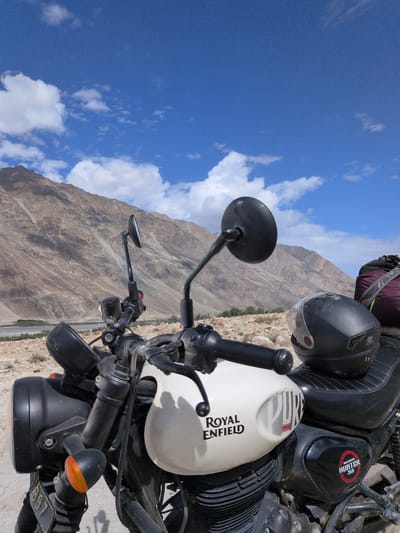 A motorcycle, identified by its "Royal Enfield HUNTER 350" badging, is parked on a rocky, arid terrain under a bright blue sky with scattered clouds. The motorcycle, primarily white and black, is positioned in the foreground, angled towards the viewer's right. It features a prominent headlight, handlebars with mirrors, a fuel tank with "ROYAL ENFIELD" and "PURE" branding, and a black leather seat. A black helmet rests on the seat. In the background, rugged, light-brown mountains rise steeply, their slopes marked with various textures and shadows. A dry riverbed or valley can be seen at the base of the mountains, with sparse vegetation. The overall impression is a solitary scene in a remote, mountainous landscape, likely within the region of Sasoma, India, given the context provided. No people are visible in the image.