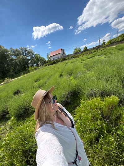 This media file captures an outdoor scene in Gay-Kodzor, Russia, on a bright, sunny day. The image is composed with a wide expanse of vibrant green vegetation dominating the lower and central parts of the frame, possibly a field of young crops like lavender. The sky is a clear, brilliant blue with scattered white, fluffy clouds, indicative of good weather.
A person is visible on the right side of the frame, from the chest up, with their head tilted slightly upwards. They have long, blonde hair and are wearing a light-colored, possibly white, textured top or dress. A wide-brimmed straw hat covers part of their head, and sunglasses are worn either on their face or perched on their head. A necklace with small white beads is visible around their neck, and a strap, possibly from a bag or camera, with pink and silver charms, crosses their chest.
In the mid-ground and background, on the left side, there are several large green trees and a visible wooden fence line. Further back, a house with a distinct red-tiled roof can be seen amidst more greenery. Utility poles with overhead wires are also present, running across the landscape. The overall atmosphere suggests a peaceful, leisurely moment spent outdoors in a rural or semi-rural setting, enjoying the sunshine. There is no visible text in the image.