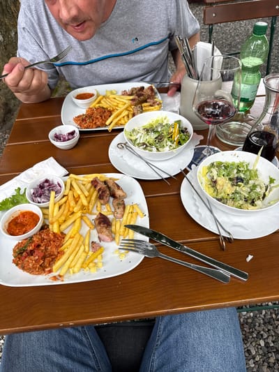 This is a top-down, medium shot of a meal being served at an outdoor restaurant. Two people are visible, one in the foreground on the left, wearing a grey t-shirt, and another on the right, whose blue jeans are visible. They are seated at a wooden table.

On the table are two plates of food, each containing grilled meat, french fries, and a tomato-based rice dish. There are also small bowls of chopped red onion and a red sauce, as well as a salad in a white bowl. Two glasses of red wine, a bottle of green-capped water, and a glass carafe are also on the table. Silverware, including a knife and fork, are laid out for the meal.

The scene suggests a meal in progress, likely at lunchtime or early evening, given the natural light. The setting appears to be casual and outdoors, possibly a beer garden or restaurant patio, with gravel visible beyond the table. There are no strong indicators of emotion, but the focus on the food and drinks suggests a relaxed dining experience. There is some German text visible on a coaster or napkin underneath one of the salad bowls.