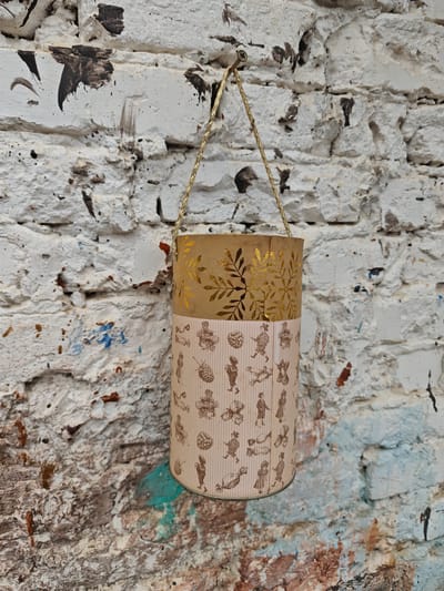A two-tiered decorative cylinder hangs from a gold chain on a heavily textured, white brick wall. The top tier is brown with gold leaf designs. The bottom tier is light pink with brown pinstripes and illustrations of people, candies, and berries. The wall has splatters of brown, black, and blue paint. The cylinder is positioned approximately in the center of the frame. A nail is visible near where the chain is attached to the wall.
The location is посёлок совхоза "Буденновец", Russia.