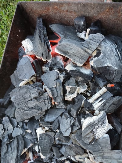 A top-down, close-up shot shows a pile of charcoal embers glowing with orange and red heat in a rusty metal brazier. The brazier is situated outdoors, with hints of green grass visible in the upper left corner. The charcoal pieces are varied in size and shape, some are black and charred, while others show white ash and streaks of red from the heat. The focus is on the texture and composition of the burning charcoal.