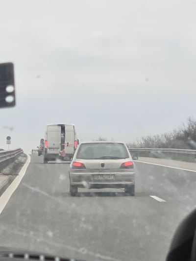 The image displays a daytime scene on a multi-lane highway under an overcast sky. The view is captured from inside a vehicle, looking forward through a windshield which shows minor imperfections and reflections.
In the mid-ground, a silver or light-beige Peugeot 306 hatchback is visible, traveling in the same direction as the camera vehicle. Its brake lights are illuminated red. The rear license plate, "PB 1523 MT," is clearly legible, indicating registration in the Plovdiv region of Bulgaria.
Further ahead in the background, a white panel van is observed, also traveling in the same direction. Its rear doors are partially open or missing, revealing a vertically mounted object, possibly a bicycle or a ladder, secured within the cargo area. Red taillights are visible on the van. Other indistinct vehicles are visible further down the road.
The highway features at least two lanes of travel in the depicted direction, separated by a broken white line. A solid white line marks the left edge of the roadway, bordering a concrete barrier or guardrail. A circular road sign, partially obscured, is mounted on the left side of the road near the barrier. On the right side of the highway, distant bare trees or shrubs are visible against the horizon. The specific location within Opalchenets, Bulgaria, cannot be definitively identified from the visual evidence, but the license plate confirms the scene is within Bulgaria.