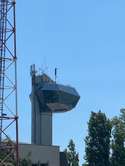 The image captures a specific architectural structure and communication infrastructure against a clear, bright blue sky in Drobeta-Turnu Severin, Romania. On the left side of the frame, a segment of a large metal lattice tower, painted in alternating red and white sections, is partially visible, extending beyond the top and bottom edges. To the right of the tower, a distinctive multi-story concrete building, known as the Water Castle of Drobeta-Turnu Severin, dominates the mid-ground. This building features a unique, dark glass observation deck or architectural element near its summit, which is multifaceted and extends outwards from the main concrete shaft. Below this glass structure, faint vertical blue lettering on the concrete column reads "PORȚILE DE FIER". The flat roof of the concrete section above the glass feature is equipped with numerous communication devices, including multiple cellular antennae, small satellite dishes, and other transmitting equipment. In the lower right quadrant, several tall, slender deciduous trees with dense green foliage are prominent. A small portion of a light-colored building is visible at the bottom left. The sky is uniformly clear and light blue, devoid of clouds, with two very small dark specks in the upper right quadrant. No individuals, animals, or specific actions are depicted within the scene.