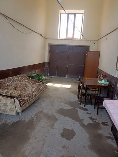 The image depicts an interior space, possibly a barn or a storage area, in the village of Aygezard, Armenia. The room is sparsely furnished and appears to be in a state of disrepair or disuse.

In the foreground, a worn sofa with a patterned fabric is positioned against the left wall. Next to the sofa, a potted plant with large green leaves is visible. The floor is concrete and wet in several areas, with dark, irregular patches indicating standing water or dampness.

Towards the center of the room, a wooden dining table is set with four chairs. On the table, there are a few items, including a vase with red flowers, a cup, and what appears to be a small device. A tall, brown wooden cabinet stands behind the table.

A large, double metal door is visible in the background, suggesting an exit or entrance to the space. Above the door, a tall, rectangular window with wooden frames allows natural light to enter, casting distinct shadows on the floor.

The walls are painted a light, neutral color, but show signs of wear and tear, with some darker staining near the floor. Electrical wiring is visible along the walls and ceiling, appearing somewhat haphazard.

The overall impression is one of a functional but neglected space, with elements of domesticity (sofa, dining set) juxtaposed with the utilitarian nature of the building.