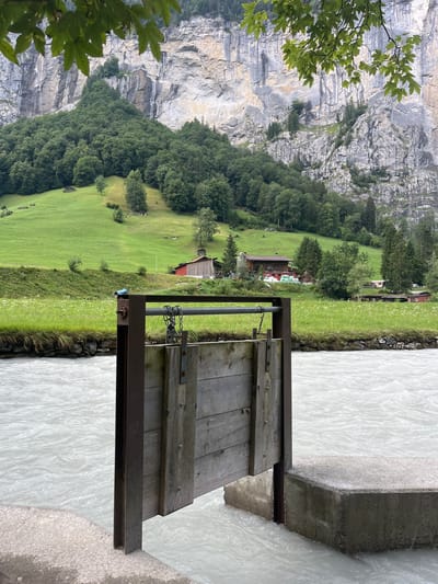 The image depicts a section of the Wengernalp railway line in Wengen, Switzerland, featuring a wooden gate mechanism situated at the edge of a fast-flowing, milky-white river. The gate, constructed from weathered wooden planks and supported by a metal frame, is partially open, allowing the river to flow through. In the background, lush green meadows ascend towards a dramatic, sheer cliff face, dotted with trees and small chalets. The scene is framed by the dappled leaves of a tree in the foreground, suggesting a natural, mountainous environment. The overall impression is one of a picturesque Alpine village setting with functional infrastructure integrated into the landscape.
