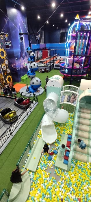 An indoor playground features a large ball pit filled with yellow and blue balls, surrounded by green platforms. Kids play within the ball pit. Tunnels and slides create a fun, interactive space. Above, soccer balls hang from the ceiling. A colorful, multi-level play structure adds to the vibrant atmosphere. The flooring displays a space-themed design with planets and gears.