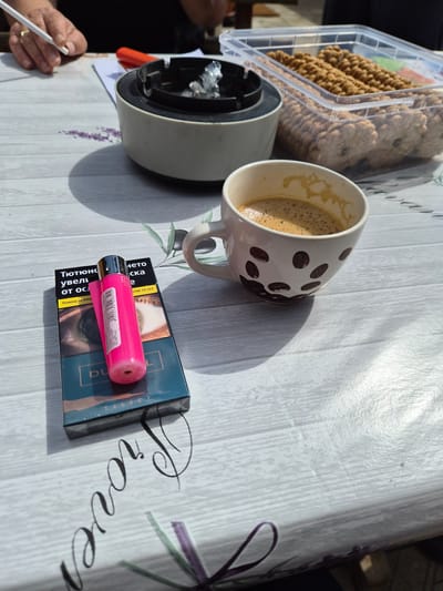 A partially consumed cup of coffee in a white mug with brown coffee bean designs sits on a light-colored tabletop with a "Proven" pattern. Beside the mug, there is an ashtray with cigarette butts. A pack of "Du Select" cigarettes with a pink lighter on top is also visible. A container of cookies is present near the mug. In the lower-left corner, a hand holding a cigarette is visible. The scene appears to be outdoors in daylight. The location is potentially Stroevo, Bulgaria, based on the "Тютюнс" text on the cigarette pack.
