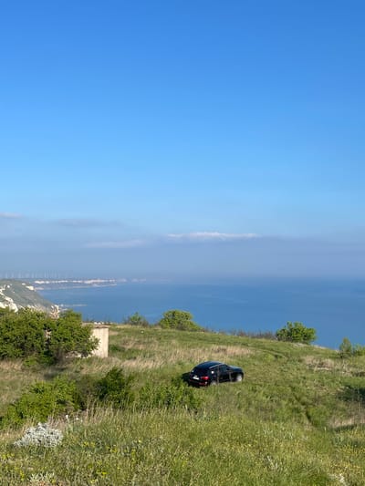 A landscape photograph captured in Bozhurets, Bulgaria, showcases a black car parked on a grassy, inclined terrain overlooking the Black Sea. The sky occupies the left side of the frame, transitioning from deep blue at the top to a hazy, lighter blue towards the horizon. The sea stretches horizontally, with a bridge-like structure visible in the distance through the haze. On the right, the green hillside is dotted with shrubs and vegetation, leading down to a cliffside with white rocky formations and more dense greenery near the bottom. A small, concrete structure is also seen among the foliage on the hillside. The car is parked diagonally on the slope, appearing to be on a rough, unpaved road.