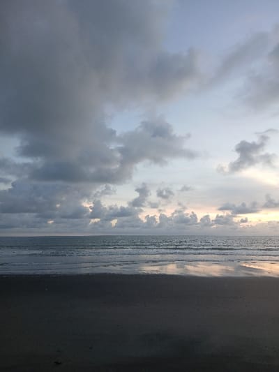 The scene depicts a beachfront environment in Tibubiyu, Indonesia, under a predominantly cloudy sky during either dawn or dusk. The upper two-thirds of the image are occupied by a sky densely covered with dark grey stratocumulus clouds, particularly concentrated in the upper left quadrant. Towards the horizon on the right, the clouds are lighter and more broken, revealing faint hues of pale orange, pink, and yellow indicating a low sun. The middle third of the image shows the ocean, characterized by calm, undulating water with several distinct lines of small waves approaching the shore. The ocean surface reflects the subdued sky light. The bottom third of the image consists of a dark, wet sandy beach. The sand reflects the overhead sky, creating a smooth, reflective surface along the waterline. No individuals, animals, structures, or identifiable objects are present within the frame.