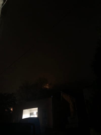 The scene depicts a residential area in ქვედა სამება, Georgia, at night under low light conditions. The sky is largely dark and overcast, with a faint, diffuse orange glow visible in the upper background, likely from a distant light source reflecting off clouds or fog. Two dark power lines extend diagonally across the upper portion of the frame. In the midground, a light-colored, single-story building is visible, featuring a brightly illuminated interior seen through a large, rectangular opening on its facade. The interior appears to have white walls and an overhead light source. To the immediate right of this building, the dark silhouette of a piece of heavy equipment, specifically an excavator arm and bucket, is partially visible, parked in an upright position. Dark, indistinct foliage and trees are present in the background, behind the building and equipment. A dark, indeterminate object, possibly the roof of a vehicle, occupies the lower left foreground. No individuals are present.