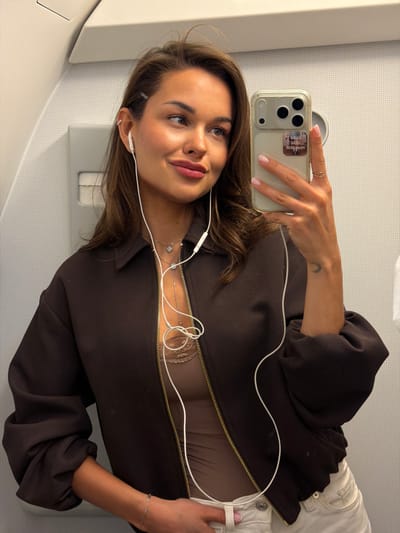 The image shows a woman taking a selfie inside what appears to be an airplane. The background shows elements typical of an airplane cabin, such as overhead storage compartments and textured wall panels. The woman is holding a phone in her right hand, using it to capture the image. She is wearing a dark brown zip-up jacket, with the zipper partially open, revealing a light-colored top underneath. She is also wearing white pants. She has earphones connected to her phone. The phone case has stickers on it. The woman has light brown hair and is wearing jewelry, including rings and necklaces. There is a bracelet visible on her left wrist. The lighting is bright, suggesting daylight conditions outside. This is inside an airplane. The location is given as Emelyanovo Airport in Russia.