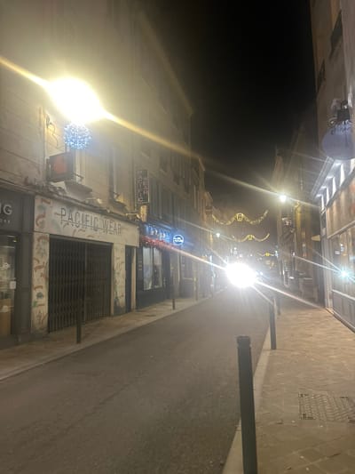 A street is illuminated at night, featuring store fronts with signs reading "Pacific Wear" and "Bar Paoli". Above, decorative lights stretch between buildings, and Christmas decorations are visible. The street surface appears wet, reflecting light from the overhead sources. A tall pole is also visible on the right. The scene is captured with a strong perspective, looking upwards towards the end of the street.