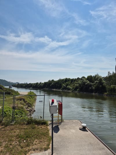 This media file depicts a scenic view of a canal or river in Passau, Germany. The sky is a vibrant blue with wispy white clouds, suggesting a pleasant daytime with fair weather. The left side of the image is dominated by the sky and the vast expanse of water, which reflects the light and shows gentle ripples. Along the right bank of the waterway, a dense line of lush green trees creates a natural border, with a concrete embankment or walkway in the foreground.
On the embankment, several objects are visible, including a white bollard attached to the concrete, likely used for mooring boats. Further along, there is a railing and a structure containing a red object and a box with a red light, possibly signaling equipment for navigation or a warning light. The area beside the embankment is covered in green grass, some of which appears dry and sparse, indicating a sunny season. A metal fence runs along the edge of the embankment, separating it from the grassy area.
In the distance, the far bank of the river is lined with more trees and hints of buildings and hills, suggesting a populated area or town. A boat is visible on the water in the far distance, adding a sense of scale and activity to the tranquil scene. The overall impression is one of natural beauty combined with the infrastructure of a working waterway. The time of day appears to be late morning or afternoon, given the bright sunlight and the shadows cast by the objects. No people are clearly visible in the image.
