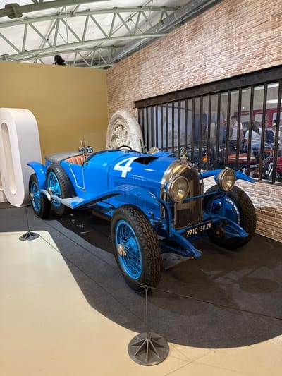 A vintage blue race car with the number 4 displayed on the side. The car has large, spoked wheels, open-wheel design, and classic headlamps. It is positioned on a grey platform within a museum setting. A barrier wall and overhead structure with brick design are visible. A display case and support beam are also featured in the background.