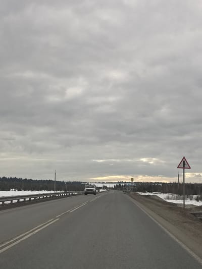 The image depicts a straight two-lane asphalt highway, likely leading into or out of Petushki, Russia, under an overcast, heavily clouded sky. The road features a double solid white line delineating the two opposing lanes. A light-colored van or SUV is visible in the distance, traveling away from the viewer in the right lane.
On the left side of the road, a silver metal guardrail runs alongside a snow-covered embankment. Beyond the embankment, a dense line of evergreen trees forms a forest, extending to the horizon. A tall, slender pole is visible behind the guardrail on the left.
On the right side of the road, in the foreground, a triangular warning sign with a red border is present, depicting a T-intersection where a minor road joins from the right. The road shoulder on the right is unpaved, showing patches of dirt and residual snow. Further down the right side, another indistinct yellow rectangular sign is visible. The distant horizon reveals a faint strip of brighter sky beneath the clouds. No people are visible. The environment indicates late winter or early spring conditions.