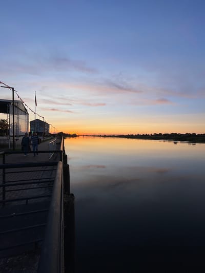 A low-angle, vertical shot captures a tranquil sunset over the Neman River in Sovetsk, Russia. A paved walkway lines the riverbank, with a dark metal railing in the foreground. Two individuals, a man and a woman, stroll along the walkway, their backs to the camera. To the left, a large outdoor stage structure with a white fabric backdrop is visible. Across the river, a treeline punctuated by buildings and a distant bridge stretches towards the horizon, where the sun dips below, casting a warm orange glow. The sky is a gradient of soft blues and pinks, with wispy clouds reflecting on the calm river surface.