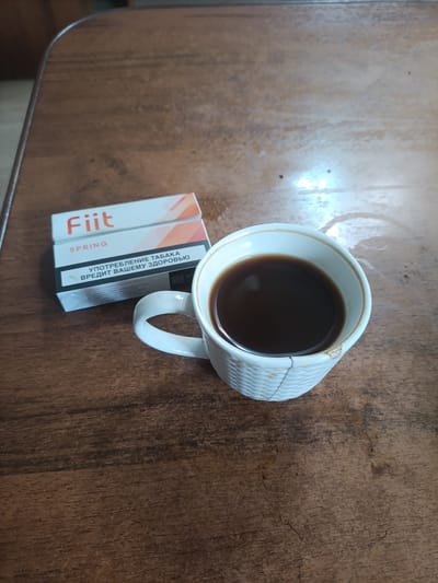 A pack of "Fiit SPRING" cigarettes and a white ceramic mug filled with dark liquid, likely coffee, are placed on a dark wooden table. The cigarette pack is partially visible, showing the brand name "Fiit" and "SPRING" in English, along with text in Russian that translates to "tobacco consumption is harmful to your health." The mug has a textured surface and a crack is visible on its right side. The scene is captured from a top-down perspective. The location is the city of Posyolok Sovkhoza "Budennovets", Russia.