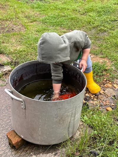 A young child, wearing a green hooded sweatshirt and grey pants, with bright yellow rain boots, is bending over a large, metal pot filled with murky water. The child's arm is submerged in the water, reaching for a red object. The pot is resting on a wet, paved surface, with a single red brick propping up one side. The surrounding environment is grassy with patches of soil and small stones. The scene appears to be outdoors, likely in a backyard or garden area. The location is in Verkhnyaya Uryakh, Russia, given the context.