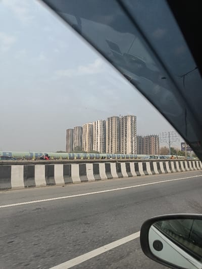 The image captures a view from inside a vehicle driving on a multi-lane highway or expressway under a hazy, overcast sky. In the foreground, the asphalt road features white lane markings. A series of black and white striped concrete barriers separate the active road from a raised median or adjacent structure.
Behind these barriers, a long, light blue or green cylindrical pipeline or covered structure runs horizontally across the mid-ground. Prominently displayed in yellow letters on this structure is the text "UTTAR PRADESH," indicating the geographical location as the Indian state of Uttar Pradesh. A few individuals are visible on or near this structure; one person in a red top and another in a white top can be discerned.
In the background, a cluster of numerous tall, multi-story residential or commercial buildings, appearing to be under construction or recently completed, dominates the skyline. These buildings are uniformly beige or light grey, with many visible windows and structural elements. To the far right, part of a billboard structure is visible, with partially obscured text that seems to include "SI" or "SIA" and "ORP".
The perspective is from the passenger side, with a dark, diagonal obstruction in the top portion of the frame, likely the car's window frame or sun visor. A side-view mirror of the car is visible in the bottom right corner, reflecting a portion of the car's body. The scene depicts an urban or semi-urban environment consistent with Ghaziabad, India, characterized by significant infrastructure and ongoing development during daylight hours.
