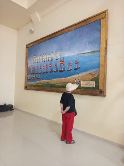 A woman wearing a white hat, black top, and red pants stands with her back to the viewer, looking at a large painting on a cream-colored wall. The painting depicts a naval battle with numerous sailing ships in a blue body of water. The ships are arranged in formations, and many fly red and yellow flags, suggesting a historical Spanish naval engagement. A sandy shore with greenery and a small settlement is visible in the foreground of the painting. A scroll with writing is also integrated into the artwork. To the left of the painting, on the floor, is a dark, curved object, possibly an old cannon. The floor is tiled with large, light-colored squares. A white speaker is mounted on the wall above and to the left of the painting. The scene appears to be in a museum or exhibition space, potentially in Coveñas, Colombia, given the context provided.