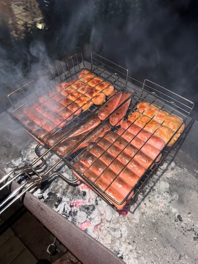 The image shows a grill basket containing sausages and patties being cooked over hot coals. The grill basket rests on the edge of a concrete fire pit. Visible in the lower left corner is smoke, presumably from the burning coals. The context of the city of Mayak, Russia is not evident in the image.
