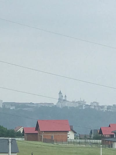 The image presents an exterior landscape view, likely captured in Șcheia, Romania, looking towards a distant cityscape atop a hill under an overcast sky. The scene is dominated by a pervasive atmospheric haze that softens details, particularly in the midground and background.

In the foreground, a verdant green field occupies the lower portion, showing signs of cultivation or regular maintenance. Scattered across this field are several residential structures, characterized by gabled red or orange-tiled roofs. A prominent, partially built or rustic-style house with a red roof and brick-like lower walls stands in the left-center, featuring a smaller white-walled section to its right. To the far right, another similar house with a red roof is visible. A house with a dark gray, sloped roof is partially visible in the bottom-left corner. Utility lines, thin and dark against the lighter sky, traverse horizontally across the upper and middle portions of the image. A utility pole is partially visible on the far left. Segmented fencing, consisting of vertical posts and horizontal elements, delineate property boundaries within the mid-ground. Green foliage from trees and bushes is visible behind some of the foreground houses on the left.

The midground is dominated by a dark green, heavily forested hill that acts as a natural separator. The top edge of this hill is indistinct due to the atmospheric haze.

The background reveals a distant city, likely Suceava, situated on the crest of the aforementioned hill. A large, light-colored architectural complex, strongly resembling a monastery or a grand church, is centrally positioned on the hilltop, featuring multiple pointed spires and domed towers. This prominent structure is identifiable as the Monastery of Saint John the New of Suceava. Adjacent to and surrounding this building are various other structures, some appearing to be multi-story and modern, extending along the hilltop. The details of these distant buildings are significantly obscured by the atmospheric haze, rendering them indistinct.

The sky is uniformly light gray, indicating an overcast condition without discernible cloud formations. No individuals, animals, or specific actions are depicted within the frame.