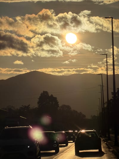The image displays a vibrant outdoor scene captured at either sunrise or sunset in Nikšić, Montenegro. The left two-thirds of the frame are dominated by a sky filled with dramatic clouds, illuminated in shades of bright white, grey, and golden-orange by the low-lying sun. A brilliant, circular sun, appearing almost white, is visible on the left side of the frame, partially obscured by these luminous clouds.

A dark, silhouetted mountain range or large hills runs horizontally across the mid-section, separating the bright sky from the darker elements on the right. Dark streetlights or utility poles with horizontal arms are silhouetted against the bright sky in the upper part of the image.

On the rightmost vertical third of the frame, a road stretches from the bottom to the top. A line of vehicles is present on this road, their headlights brightly illuminated and creating pronounced lens flares, especially from the cars closer to the bottom. These cars appear mostly as dark silhouettes against the reflective road surface. Dark trees and vegetation are visible along the road's edges, particularly in the upper right.

The scene captures a moment of transition during the day, with the sun's low angle and the active car headlights indicating either an early morning commute or late afternoon traffic. The dramatic lighting and cloud formations create a striking and atmospheric visual. No visible text is present in the image.