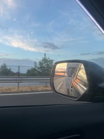 The media file presents a rotated, intimate view from inside a moving vehicle, focusing on the exterior side mirror and its surroundings. The primary subject is the car's side mirror, which appears speckled or dirty, reflecting a striking sunset or sunrise with vibrant orange and yellow hues across the sky. Reflected in the mirror, a white vehicle (possibly a van or small truck) is visible on a multi-lane road or highway, alongside a guardrail.
Outside the car window, beyond the mirror, the sky is a soft blue with scattered white clouds, suggesting a slightly different angle or illumination compared to the intense colors reflected in the mirror. Trees and distant landscape elements are faintly visible through the window, behind what appears to be a highway guardrail. The car's dark, textured interior door panel is visible on the right side of the frame.
The image captures the specific time of day during either dawn or dusk, characterized by the warm, intense colors of the low sun. The setting appears to be a journey on a highway, possibly near Makó, Hungary, given the context. There is no visible text in the image, and no specific individuals are present.