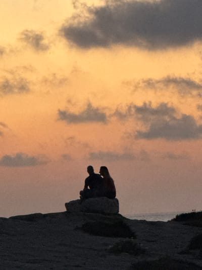 A silhouette of a person perched on a rocky cliff against a backdrop of a pastel-colored sunset sky. The sky displays a mix of soft orange and gray hues with scattered cloud formations. The cliff edge is rugged and dark, contrasting with the lighter sky. The person appears to be sitting or kneeling, enjoying the view or moment.