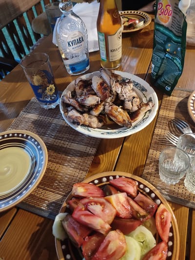 The image depicts a close-up, high-angle view of a wooden table set with food and drinks, likely outdoors in Igenche, Russia. A platter of grilled chicken wings is centrally located, accompanied by a salad of chopped tomatoes and cucumbers in a decorative bowl. To the left, a glass with a sunflower design sits next to a bottle of "Купава" vodka. To the right, a bottle of what appears to be kvass or a similar beverage, a pouch of mayonnaise, and two small shot glasses are visible. A fork rests to the right of the shot glasses. The overall scene suggests a casual meal or gathering, possibly a barbecue, given the grilled chicken and outdoor setting implied by the visible wooden planks and slightly blurred background of what appears to be a wooden bench and greenery.