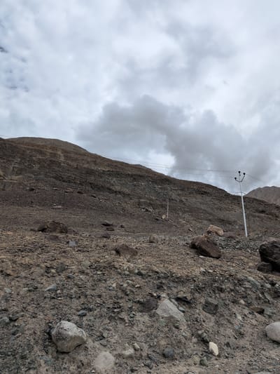 The image depicts a barren, rocky hillside with scattered boulders and a few utility poles with electrical wires. The terrain is steep and appears to be composed of loose soil and rocks. The sky is overcast with thick, grey clouds. In the foreground, various sizes of stones and pebbles cover the ground. A white utility pole with three insulators and electrical wires running across the hillside is visible on the right side of the image. Another similar pole is visible in the middle distance on the left. The scene suggests a remote, undeveloped, or mountainous area. The location is the city of Panamik, India.