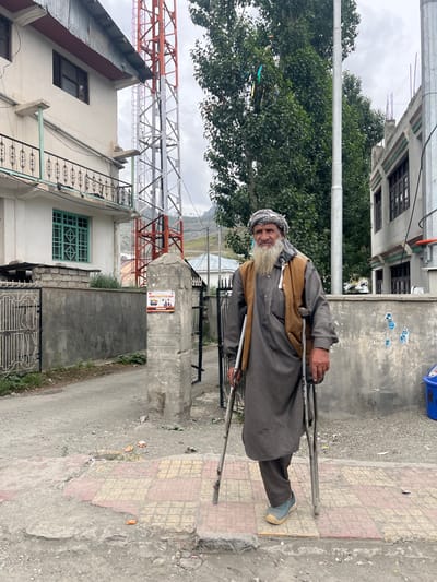 The image depicts a man with a long white beard and a checkered headdress walking with the aid of two crutches. He is wearing a grey traditional outfit and a brown vest. He is walking on a paved sidewalk with a checkered pattern of pink and yellow tiles. To his left, there is a grey concrete wall and a black metal gate. Behind him, there are buildings, including a white building with a balcony, a tall metal communication tower, and a grey building with dark windows. Lush green trees are visible behind the tower and the buildings. The ground is mostly unpaved and dusty, with scattered debris. In the background, mountains are faintly visible under an overcast sky. The scene appears to be in a town or village, likely in Dras, India, given the mention of "SABROO SURYATHON 2026" on a poster attached to a concrete pillar to the left of the man. The poster features small images of people and text. To the far right of the image, a blue garbage bin is partially visible.