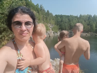 A family is enjoying a day at a quarry lake. In the foreground, a woman with dark hair and sunglasses takes a selfie, holding a young child. The child is facing away from the camera, their back visible. Behind them, a man with short hair holds another child. The man is wearing orange swim trunks, and the child he is holding is wearing a red and white patterned bikini bottom. The background features a rocky quarry with lush green trees and a clear blue sky. The sun appears to be high in the sky, suggesting it is midday. The water in the lake is dark and still. The overall atmosphere is one of leisure and family fun in a natural outdoor setting. The image was captured in Výkleky, Czechia. There is no visible text in the image.