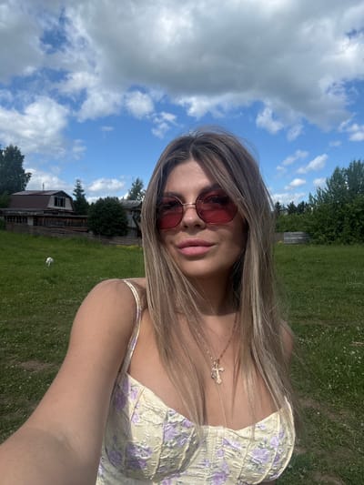 This is a portrait-oriented selfie taken outdoors. The primary subject is a young woman with long, blonde hair and pink lipstick. She is wearing red-tinted sunglasses and a light-colored dress with a floral pattern. Around her neck, she has a delicate necklace with a cross pendant.
The setting appears to be a rural area with a grassy field and trees in the background. There are also some wooden structures that resemble buildings or fences. The sky is blue with scattered white clouds, suggesting it is daytime and the weather is pleasant. The image is well-lit, indicating it was taken on a sunny day. There is no visible text in the image. The woman's pose and facial expression suggest a casual and relaxed mood. The overall impression is one of a sunny day spent outdoors in a peaceful, rural environment.