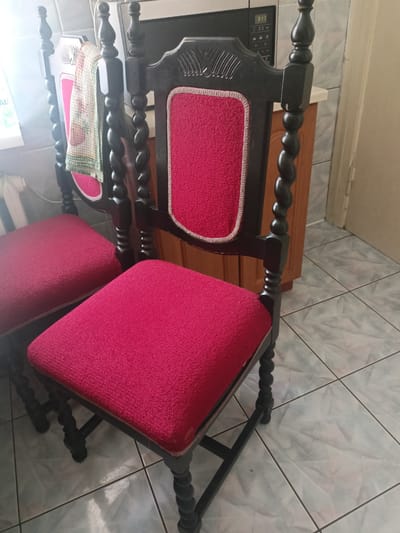 Two dark wooden chairs with ornate carved details and vibrant red upholstered seats are the main subjects of this media file. The chairs have twisted legs and detailed backs. One chair is positioned in the foreground, with its seat clearly visible and taking up a significant portion of the frame. The second chair is partially visible behind and to the left of the first, with its seat also upholstered in red. A checkered cloth, with patterns in green, white, and orange, is draped over the back of the second chair.
The chairs are set against a backdrop that suggests an indoor domestic setting, possibly a kitchen or dining area. To the left, a window with a bright light source casts a glare, suggesting it might be daytime. A black microwave with a digital display showing "3:17" is visible behind the chairs, indicating a kitchen appliance. The floor is tiled with light-colored square tiles and dark grout lines. A light-colored cabinet or counter is also partially visible. The overall impression is a casual, everyday scene within a home. There are no people visible, and no specific event is taking place beyond the static arrangement of the furniture. The time of day is likely daytime, given the light from the window. The location context suggests Grudziądz, Poland.
