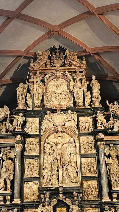 This image depicts the highly ornate High Altar, also known as the Epitaph of Archbishop Johann Schweikhard von Kronberg, located in the Collegiate Church of St. Peter and Alexander (Stiftskirche St. Peter und Alexander) in Aschaffenburg, Germany.
The altarpiece is a multi-tiered sculptural monument composed of contrasting light-colored stone, primarily for the figures and reliefs, and dark, polished marble for architectural elements such as columns, pilasters, and cornices. The structure extends vertically towards a vaulted ceiling, characterized by light grey plaster with reddish-brown ribbed arches.
At the uppermost section, a prominent central coat of arms or crest, surmounted by a cross and a crown, is flanked by two reclining winged figures (angels). Below this crest, a circular relief depicts a dense gathering of figures, including putti, in what appears to be a celestial scene. This circular relief is flanked by standing figures: on the left, a robed figure holds a tall staff topped with a cross; next to him, a winged angel. On the right, a winged angel holds an unidentified object, followed by another standing robed figure.
The middle tier features a large central arched niche containing a detailed relief of the Crucifixion. Christ is depicted on the cross, surrounded by multiple figures at its base, including draped individuals representing Mary and John, among others. This central relief is framed by dark marble columns. To the left and right of the Crucifixion, the dark marble surfaces are adorned with multiple rectangular relief panels, each depicting various biblical or devotional scenes involving multiple figures in active poses, such as gatherings, processions, or moments of interaction. Above the Crucifixion arch, a smaller central panel displays the "IHS" monogram, flanked by two kneeling angels.
Numerous smaller putti and decorative carvings are integrated throughout the structure, connecting the different tiers and panels. The figures exhibit detailed drapery and varied postures, characteristic of Baroque sculpture. The entire monument is illuminated, casting some shadows on the upper ceiling vaults.