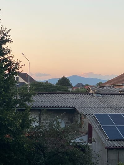 The image captures a view of rooftops and distant mountains under a dusky sky, possibly taken from a residential area in Khust, Ukraine. In the foreground, several weathered, grey tiled roofs dominate the scene, with one prominently featuring a cluster of dark blue solar panels. A large, leafy green tree partially obscures the view on the left side. Beyond the immediate rooftops, smaller houses with terracotta-colored and darker roofs are visible, interspersed with trees. In the distance, a range of blue-tinged mountains rises, partially shrouded in wispy, light-colored clouds that are catching the soft hues of the sunset. A single street lamp on a tall pole stands between the foreground and the distant landscape. The overall impression is one of a tranquil evening in a semi-urban or rural setting.