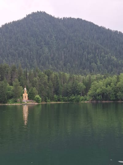 A wide shot captures a serene lake with a forested mountain in the background. In the middle ground, a small, golden-domed church stands on the water's edge, its reflection shimmering on the surface. Lush green trees surround the church and line the shore, with the dense forest continuing up the slopes of the mountain. The sky above is overcast. The location is identified as Vydrino, Russia.
