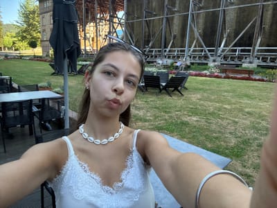 In a selfie taken at the Bad Orb graduation tower (Gradierwerk), a young woman with dark hair is the central subject. She is wearing a white camisole with lace trim and a shell necklace. Sunglasses are perched on her head. She is puckering her lips for the camera. In the background, the large wooden graduation tower structure is visible, along with tables and chairs set up outdoors. Several people are seated on benches in the distance, enjoying the green space and the historical structure. The environment appears to be a park or garden area, with manicured lawns and flower beds.