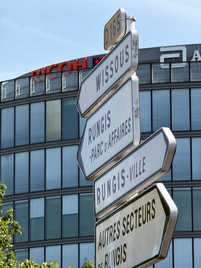 The image captures a close-up view of a directional signpost in Rungis, France. The signpost features multiple white, arrow-shaped signs with black text indicating directions to various locations. The top sign displays "D165," likely a road number. Below this, signs read "WISSOUS," "RUNGIS - PARC D'AFFAIRES," "RUNGIS-VILLE," and "AUTRES SECTEURS DE RUNGIS." In the background, a modern building with a glass facade is visible, with the red "RICOH" logo prominently displayed on its upper section. The sky above is clear and blue. The overall scene suggests an urban or semi-urban environment, likely a business district or commercial area, given the signage and the modern architecture. No people are present in the image, and the focus is on the directional signage and the surrounding architecture.