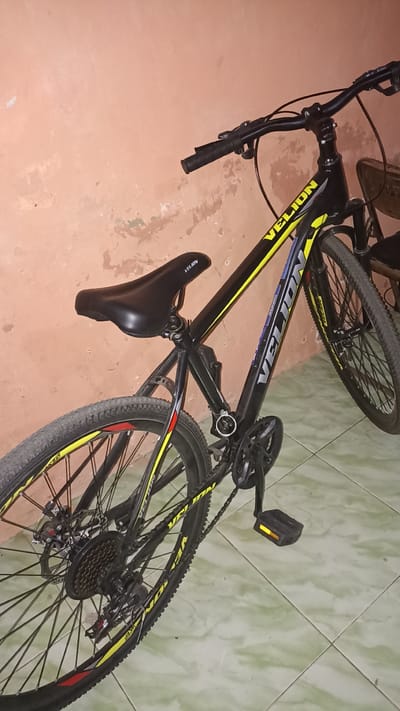 A black Velion brand bicycle is leaned against a textured peach-colored wall. The bicycle has yellow accents on the frame and wheels. The saddle is black and the handlebars are black with black grips. The bicycle has disc brakes on the rear wheel. The rear tire has a knobby tread pattern. The bicycle is positioned on a light green tiled floor. The image is taken indoors, with the floor tiles suggesting an indoor environment, potentially a garage or a room within a residential building. The background wall shows signs of wear and peeling paint. A portion of a chair or furniture is visible in the upper right corner of the image. The lighting is moderate, casting some shadows. There are no people visible in the image. The location is identified as Sedayu, Indonesia, based on the provided context, but the image itself does not contain explicit visual cues for this specific location. The image is a medium shot of the bicycle.
