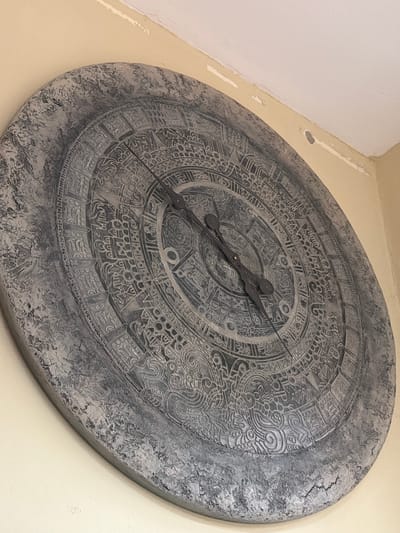 A large, circular clock, resembling a stylized Aztec calendar, is mounted on a light yellow wall. The clock face is intricately carved with various symbols and patterns, and features two dark-colored hands. The outer edge of the clock has a rough, textured, stone-like appearance. The wall has some minor imperfections and a slight shadow line near the top. The image is taken from a slightly upward angle. No people are visible in the image. The location of this clock in Maloyaroslavets, Russia cannot be determined from the image alone.