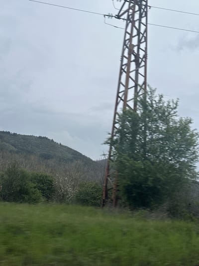 A slightly out-of-focus, horizontally-oriented photograph shows an overcast landscape. A metal power line tower is situated on the left side of the frame, with power lines strung from the top. A tree partially obscures the tower. In the background, a hillside covered in trees is visible. In the foreground, there is green vegetation. The location is possibly near Mirkovo, Bulgaria, judging from the landscape.