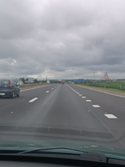 The image depicts a view from inside a vehicle traveling on a highway under a cloudy sky. The highway is paved with asphalt and has clear lane markings, including white dashes on the right and solid white lines on the left. Several vehicles are visible on the highway, including a dark-colored car in the left lane and a red vehicle further ahead in the right lane. In the distance, there are some trees and buildings. A triangular warning sign with a red border and a white interior is visible on the right side of the road, along with a blue road sign. The overall scene suggests an overcast day, likely late morning or afternoon, given the ambient light. The image appears to be taken in Belarus, specifically within the Verciiališkaŭski Sel'ski Savet region, based on the provided context. The focus is on the road and the driving experience, with no people clearly visible as subjects. The weather conditions appear to be cloudy and possibly cool, contributing to a somewhat muted color palette.