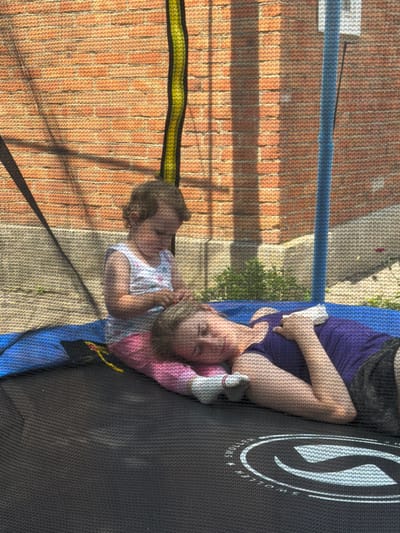 In the city of Komsomolskiy, Russia, two individuals, a younger child and an adult, are lying on a black trampoline. The trampoline has a safety net enclosure made of woven material, with visible poles. The background consists of a brick wall with sunlight casting shadows, and some greenery and a flower. The adult, lying on their back with their head resting on their arm, wears a purple top and dark shorts. The child, sitting up and leaning on the adult, is wearing a white floral-patterned top and pink bottoms. The child appears to be interacting with something in their hands. The overall scene suggests a casual, outdoor activity.