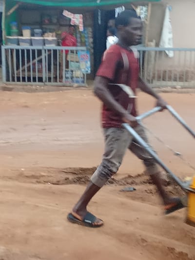 A man is seen from the side in Kagini, Nigeria, operating a piece of heavy machinery, possibly a compactor, on a dirt road. He is wearing a short-sleeved red shirt and grey pants, with black and orange sandals on his feet. The man is pushing the machine with both hands. In the background, a small market stall is visible with various items displayed, including what appear to be food items and drinks. A metal fence separates the stall from the road. The environment is dusty and appears to be undergoing construction or road maintenance.