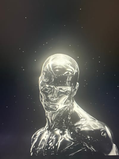 The image shows a highly reflective, metallic bust of a human-like figure against a dark background speckled with small, white dots resembling stars. The figure has sharply defined features, including a prominent jawline, nose, and eye sockets. Light emanates from behind the head, creating a bright halo effect. The overall aesthetic is futuristic and possibly extraterrestrial. There is no indication of a specific city or location within the image, and therefore no identifiable link to Vancouver, United States of America.